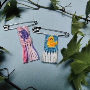 Könnte beinhalten: Zwei silberne Sicherheitsnadeln, die jeweils mit einem kleinen gewebten Wandbehang versehen sind. Eine Nadel zeigt einen rosa Hintergrund mit einem lila Dinosaurier-Motiv und rosa-lila Fransen. Die andere hat einen blauen Hintergrund mit einer gelben Ente.
