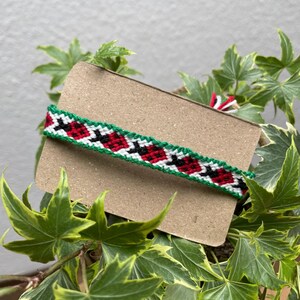 Könnte beinhalten: Ein gewebtes Freundschaftsarmband mit einem sich wiederholenden Muster aus roten Marienkäfern mit schwarzen Punkten auf weißem Grund, umrandet von dünnen grünen Linien. Das Armband ist horizontal auf einem rechteckigen hellbraunen Karton befestigt.