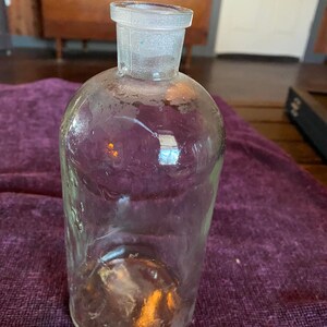 May include: A clear glass bottle with a wide mouth and a glass stopper. The bottle is empty and has a textured surface.