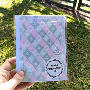 May include: A Kindle Paperwhite 11 cover in a clear plastic sleeve. The cover features a pastel diamond pattern in pink, blue, and white. A circular sticker on the cover reads "Kindle Paperwhite 11". The background is a grassy area with a wooden fence.