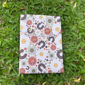 May include: A white fabric envelope with a flap closure and a pocket on the front. The envelope is decorated with a repeating pattern of brown, orange, and yellow circles and crescent shapes. The envelope has two gold-coloured metal snaps.
