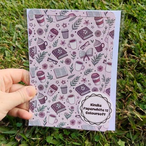 Peut inclure: Une carte à motifs avec un motif répété de livres, de tasses à café, de cupcakes, de fleurs et de lunettes dans des tons de violet et de blanc. La carte est dans une pochette en plastique transparent. L'autocollant sur la carte indique "Kindle Paperwhite 12 Coloursoft".