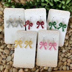 May include: Six pairs of glitter bow earrings on white cards. The earrings are silver, red, green, gold, and pink.