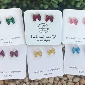 May include: Six pairs of glitter bow earrings on white cards. The earrings are in various colors, including green, red, pink, gold, and blue. The cards have the text "Stainless Steel - Hypoallergenic - Nickel Free" and "hand made with love in michigan".