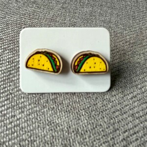 May include: A pair of small, wooden earrings shaped like tacos. The tacos are yellow with green lettuce and brown meat fillings.