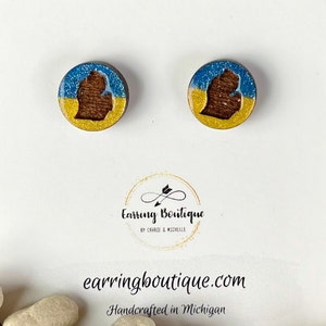 May include: A pair of stud earrings featuring a blue, yellow, and brown Michigan state outline. The earrings are handcrafted in Michigan.