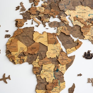 May include: A wooden world map with detailed country outlines and names, including "ARKTISCHER OZEAN" at the top. The map features various shades of brown and tan, with small wooden airplane and ship accents. The map is mounted on a white surface.
