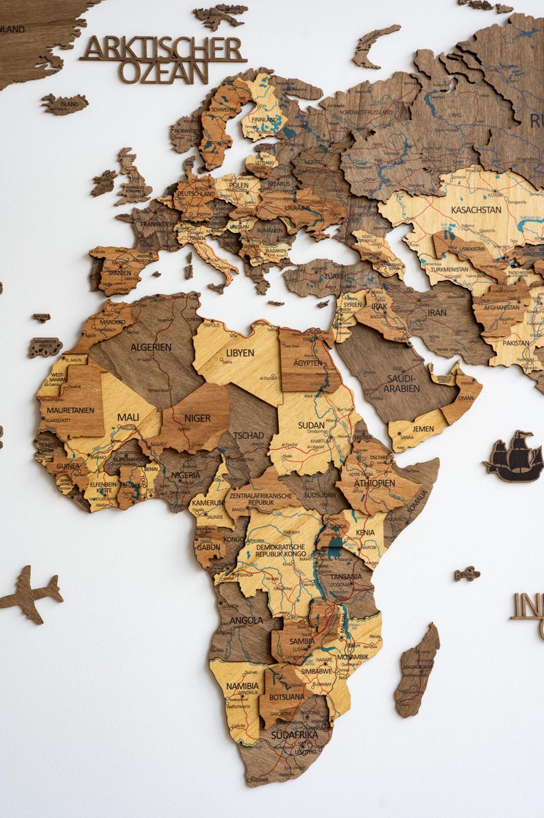 May include: A wooden world map with detailed country outlines and names, including "ARKTISCHER OZEAN" at the top. The map features various shades of brown and tan, with small wooden airplane and ship accents. The map is mounted on a white surface.