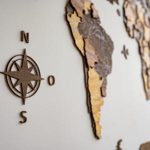 May include: Wooden wall art featuring a compass rose and a world map. The compass rose is in the center, with the letters N, S, W, and O around it. The world map is in the background, with continents in shades of brown and gold.