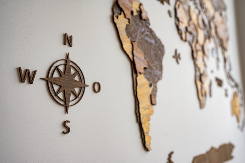 May include: Wooden wall art featuring a compass rose and a world map. The compass rose is in the center, with the letters N, S, W, and O around it. The world map is in the background, with continents in shades of brown and gold.