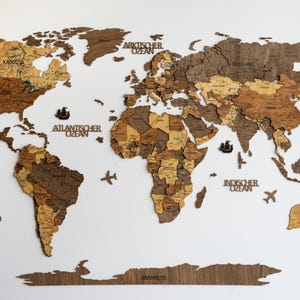 Könnte beinhalten: Eine Weltkarte aus Holz mit detaillierten Länderumrissen in Braun- und Beigetönen, montiert auf weißem Hintergrund. Enthält eine Windrose, Schiffe und Flugzeugausschnitte. Text kennzeichnet Ozeane und Kontinente.