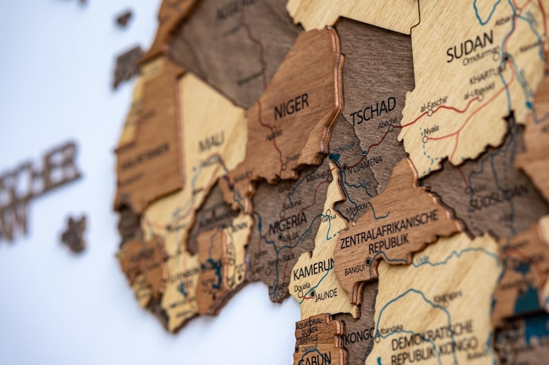 May include: A detailed wooden map of Africa, featuring countries like Nigeria, Niger, and Chad. The map is crafted from various shades of brown wood, with country names and geographical details etched into the surface. The map is mounted on a white background.