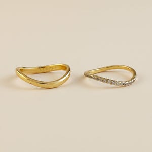 May include: Two gold rings with a wavy, curved band design. The ring on the left is a plain, polished gold band, while the ring on the right features a row of small, clear, sparkling stones set along the top edge of the curved gold band.