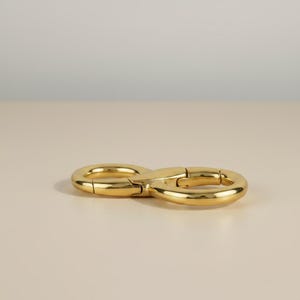 May include: A gold-colored, infinity-shaped clasp. The clasp is made of a shiny, metallic material and has a smooth surface. The clasp is designed to open and close, with a hinged mechanism. The background is a neutral beige.