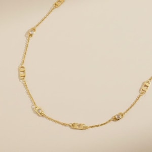 Puede incluir: Un collar de cadena en tono dorado que presenta una serie de eslabones ovalados alargados que se repiten, intercalados con pequeñas piedras redondas transparentes engastadas en marcos metálicos circulares, creando un patrón simétrico.