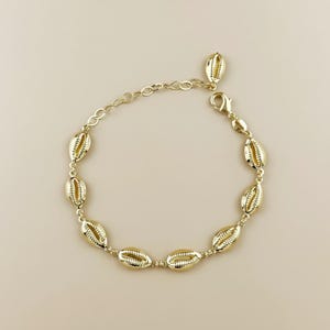 May include: Gold-toned bracelet featuring multiple seashell charms. The bracelet has a delicate chain and a lobster clasp closure. The seashells are detailed with textured ridges and are evenly spaced along the chain. The jewelry is displayed on a neutral background.