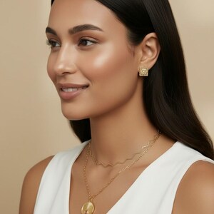 May include: A gold-toned jewelry set consisting of square stud earrings with a textured border and a layered necklace. The necklace features a delicate zigzag chain and a longer chain with a round pendant displaying a sunburst design.
