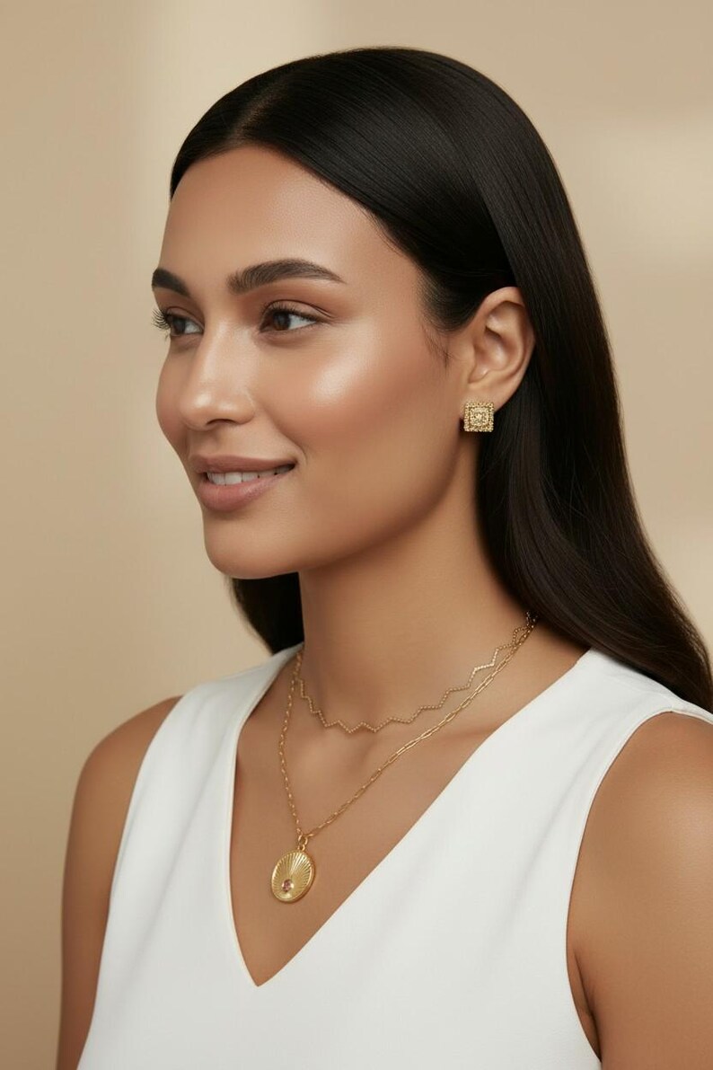 May include: A gold-toned jewelry set consisting of square stud earrings with a textured border and a layered necklace. The necklace features a delicate zigzag chain and a longer chain with a round pendant displaying a sunburst design.