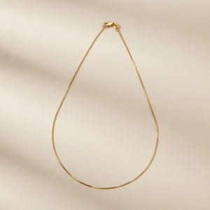 May include: A delicate gold-colored chain necklace. The necklace is a simple, elegant design, with a fine chain and a small clasp. The necklace is displayed on a neutral background, highlighting its minimalist style. A perfect accessory for everyday wear.