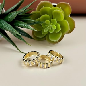 May include: Three gold-colored rings with rectangular, clear gemstones set around the band. The rings are displayed on a light-colored surface, with green plants in the background. The rings are a jewelry item.