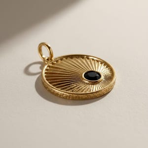 May include: A circular gold pendant featuring a textured outer rim and a sunburst pattern radiating from a single, round, black gemstone set off-center. The pendant includes a small, circular jump ring at the top for attachment to a chain.