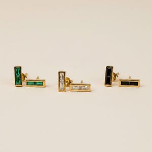 May include: Three pairs of gold-tone stud earrings, each featuring a rectangular bar design. One pair contains green baguette-cut stones, one pair contains clear white baguette-cut stones, and one pair contains black baguette-cut stones.