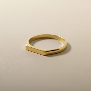 May include: A gold-colored ring with a rectangular top and a smooth, polished finish. The ring has a minimalist design and is set against a neutral background. The word "JOVTI" is engraved on the inside of the ring.