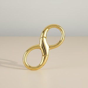 May include: A gold-colored metal infinity-shaped clasp. The clasp has two rounded loops connected by a central bar. The object is made of shiny, polished metal and is set against a neutral background.