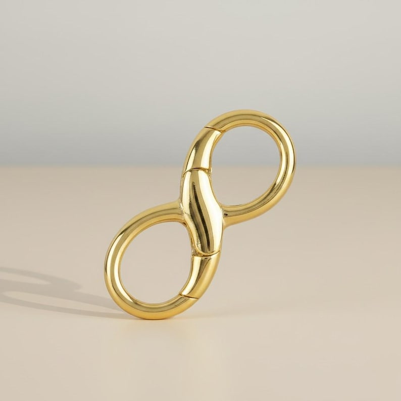 May include: A gold-colored metal infinity-shaped clasp. The clasp has two rounded loops connected by a central bar. The object is made of shiny, polished metal and is set against a neutral background.