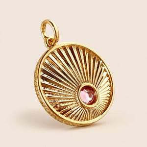 May include: A circular gold pendant featuring a sunburst pattern of radiating lines and a single round pink gemstone set off-center. The pendant includes a small gold jump ring at the top for attachment to a chain or cord, with a textured edge.