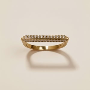 May include: A gold-colored ring with a rectangular top encrusted with small, clear gemstones. The ring has a simple band and a geometric design, suitable for everyday wear. The ring is shown on a neutral background.