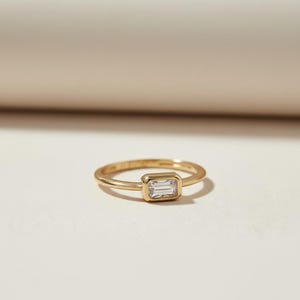 May include: A gold ring with a rectangular, clear gemstone. The ring band is thin and the gemstone is set in a rectangular bezel. The ring is displayed on a light-colored surface.