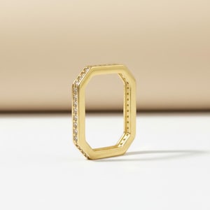 May include: A gold-toned ring with an octagonal shape, featuring a flat band design. The outer edges of the ring are lined with a series of small, clear, sparkling stones set along the perimeter, creating a continuous, reflective, and geometric look.