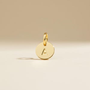Könnte beinhalten: Ein goldfarbener, kreisförmiger Anhänger mit dem Buchstaben "A" in der Mitte eingraviert. Der Anhänger hat oben eine kleine Schlaufe zum Befestigen an einer Kette oder einem Armband. Der Hintergrund ist einfarbig und neutral.