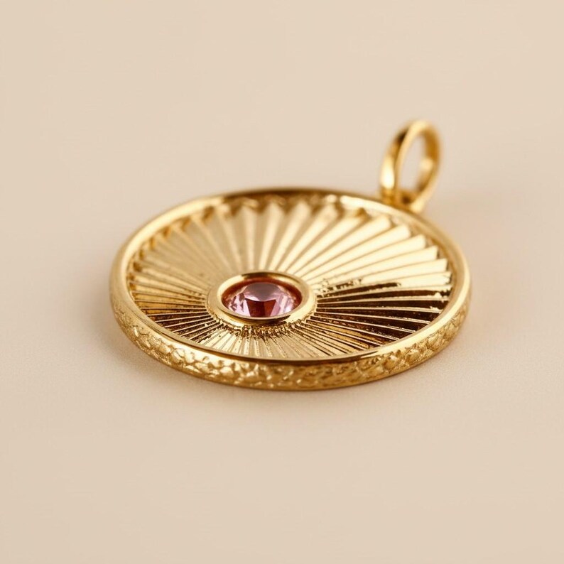 May include: A circular gold pendant featuring a sunburst pattern radiating from a central, round, light pink gemstone. The pendant has a textured outer edge and a small, smooth gold loop at the top for attachment to a chain or necklace.