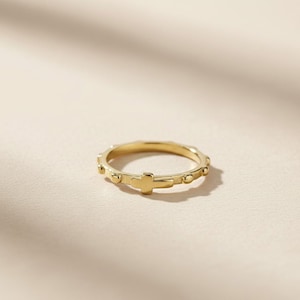 May include: A gold-colored ring with a cross and small bead details. The ring is circular and sits on a light-colored surface. The ring's design features a cross in the center and small beads around the band.