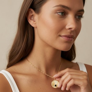 May include: A gold-toned necklace featuring a circular pendant with a sunburst-inspired engraved pattern and a small, centered black stone. The pendant hangs from a delicate, elongated oval link chain that rests against the collarbone area.