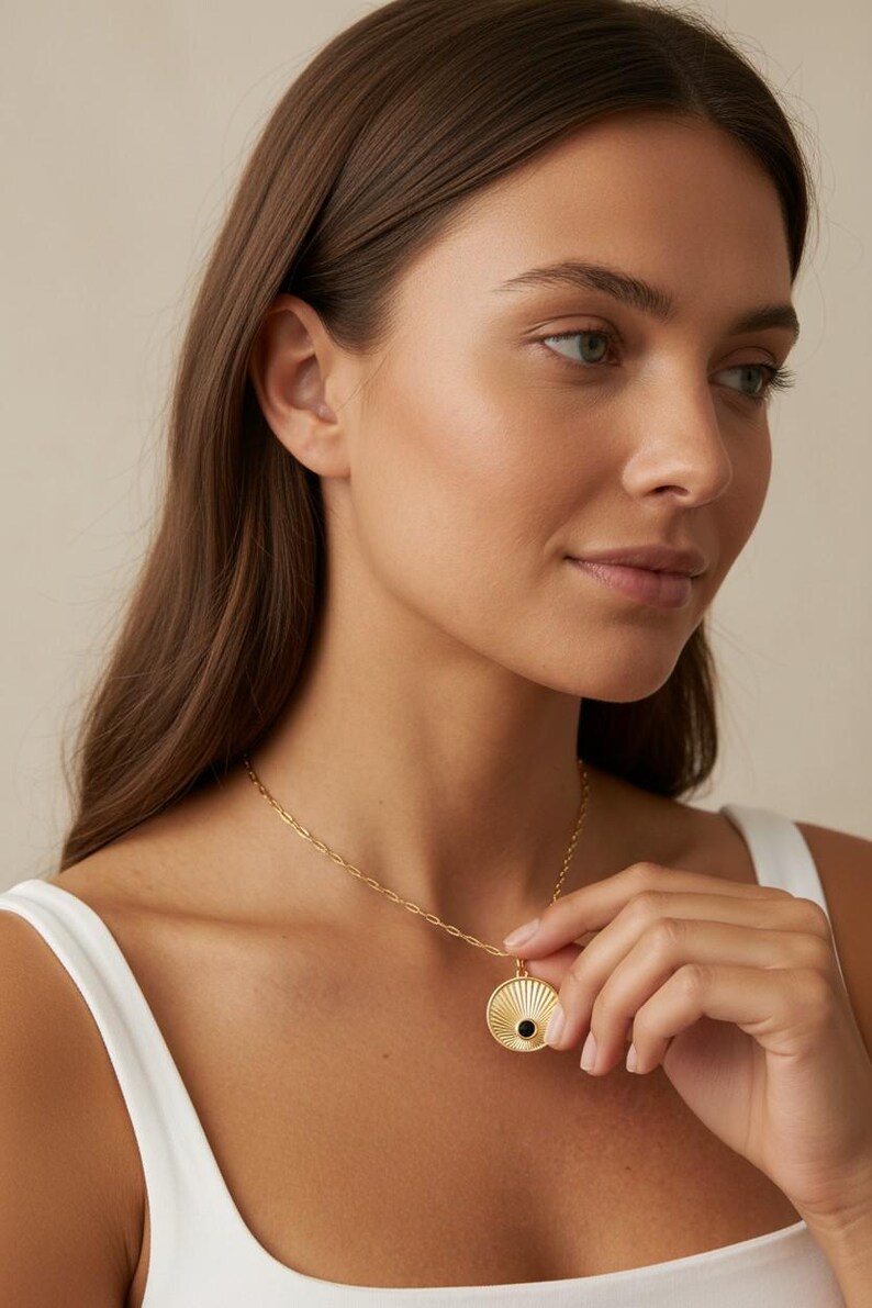 May include: A gold-toned necklace featuring a circular pendant with a sunburst-inspired engraved pattern and a small, centered black stone. The pendant hangs from a delicate, elongated oval link chain that rests against the collarbone area.