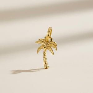 May include: A gold-colored palm tree charm with a detailed design. The charm features a loop at the top for attachment and a textured trunk and fronds. The charm is likely made of metal and is designed to be a pendant or charm.