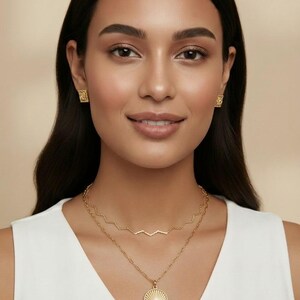 May include: A gold jewelry set consisting of a pair of small square stud earrings and a two-layer necklace. The top layer is a thin zigzag chain, while the bottom layer features a round pendant with a sunburst design and a central stone.