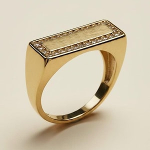May include: A gold signet ring featuring a rectangular top with a brushed matte finish. The rectangular centre is framed by a border of small, clear, round-cut stones set into the gold band, creating a polished and structured appearance.