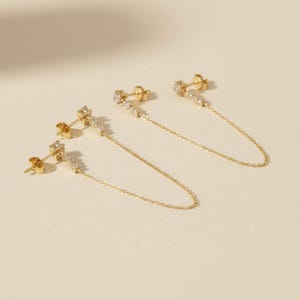 May include: A pair of gold chain earrings featuring a stud component with a rectangular bar encrusted with small clear stones. A delicate gold chain connects the stud to a second post, creating a draped effect with a polished metallic finish.