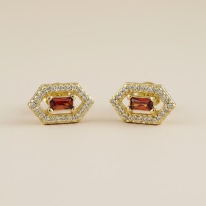 May include: A pair of gold stud earrings featuring a hexagonal frame encrusted with small clear white stones. At the center of each hexagon is a single rectangular red gemstone set horizontally, creating a geometric and layered appearance.