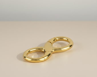 May include: A gold-colored infinity-shaped clasp. The clasp is made of a shiny, smooth metal and is resting on a light beige surface. The design is simple and elegant, with rounded edges.
