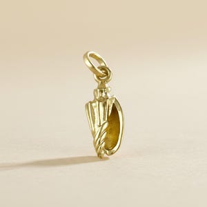 Puede incluir: Un colgante dorado con forma de concha. El dije tiene un diseño de cuerda trenzada texturizada en la parte inferior y un pequeño lazo en la parte superior para sujetarlo a un collar o pulsera. El colgante está sobre un fondo neutro.