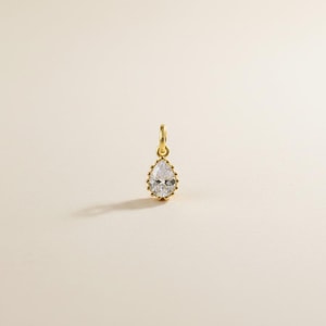 May include: A gold-colored pendant with a pear-shaped, clear gemstone. The gemstone is surrounded by a beaded gold setting and hangs from a small gold loop. The pendant is displayed on a light beige background.