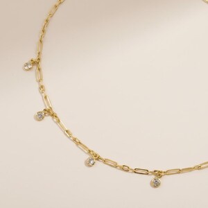 Puede incluir: Un collar dorado con un diseño de cadena delicado. El collar presenta una serie de pequeñas piedras preciosas redondas y transparentes, espaciadas uniformemente a lo largo de la cadena. La cadena tiene un diseño de eslabones tipo clip. El collar se muestra sobre un fondo neutro.