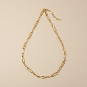 Pode incluir: Um colar dourado com uma corrente de elos retangulares alongados e elos circulares menores. O colar é exibido em um fundo neutro, destacando seu design simples, mas elegante. É uma joia.