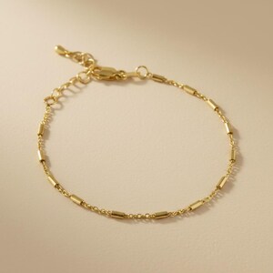 May include: A delicate gold-colored bracelet with a chain and small, cylindrical beads. The bracelet has an adjustable clasp for a comfortable fit. The jewelry is displayed on a neutral background, highlighting its simple elegance.