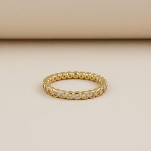 May include: A gold-colored eternity ring with a continuous row of small, clear gemstones. The ring is set against a neutral background, highlighting its polished finish and the sparkle of the stones. The ring is a classic piece of jewelry.